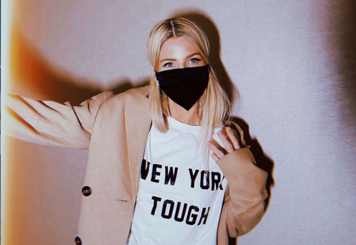 blonde woman standing front of a white wall in black silk kes mask dawning a great new york tough tshirt